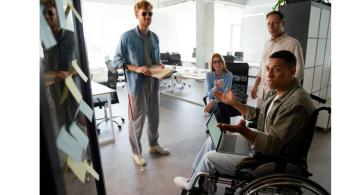 accessibilité numérique des personnes en situation de handcap : groupe de gens dont une personne en fauteuil et une personne mal voyante travaillant autour d'un ordinateur et de post-it